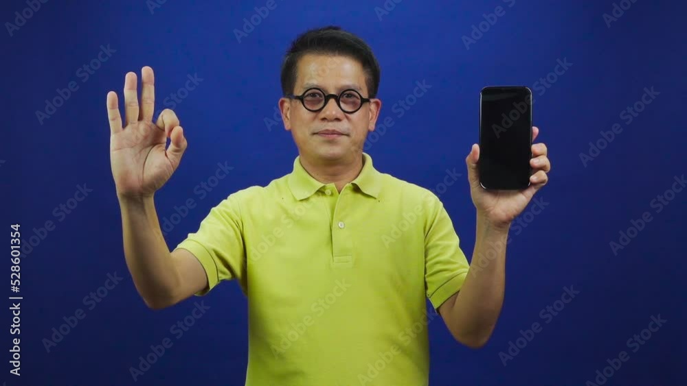 Portrait of smiling middle-aged asian man in casual yellow polo shirt showing smartphone and giving ok sign isolated on blue background in studio, looking happiness and attractive leadership