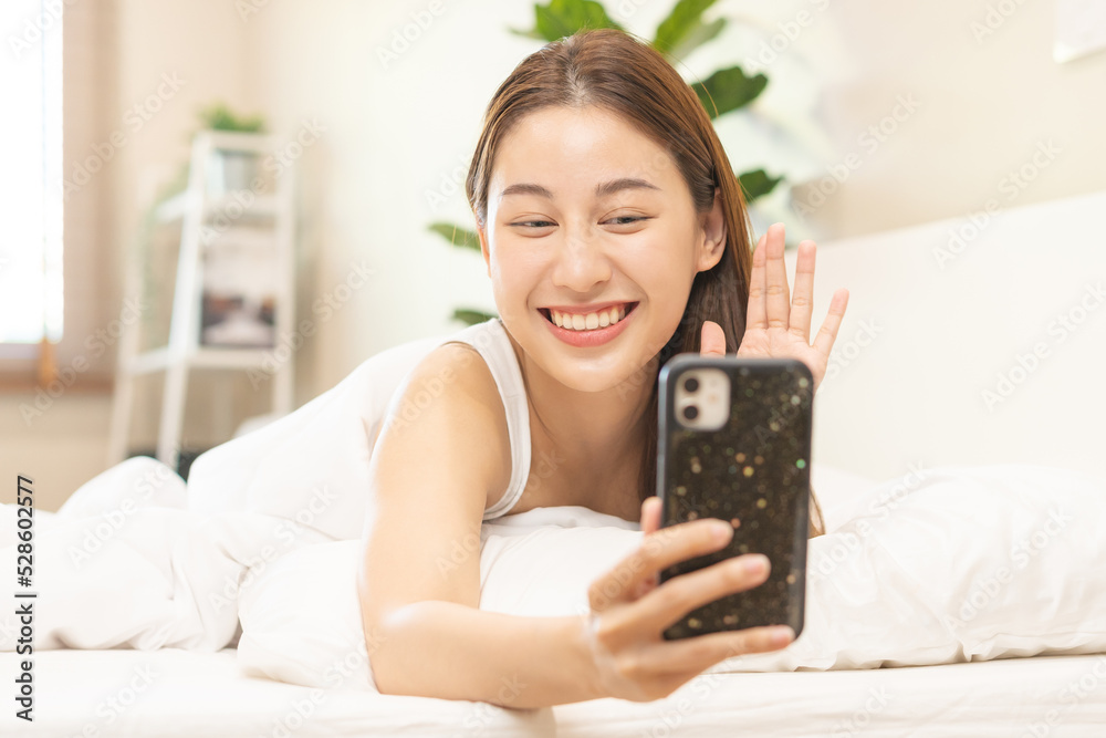 Attractive smile pretty asian young woman using and playing mobile phone, smartphone have video call or chat, lying down relax in bedroom wake up with covered blanket in cozy white bed in the morning