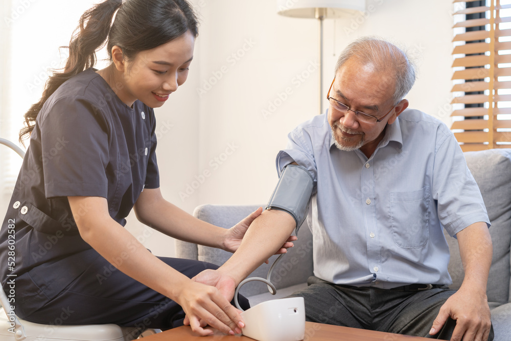 Obraz premium Pressure measurement of hypertension in elderly man sitting on couch, young woman nurse or caregiver care checking, measuring heart, blood by using sphygmomanometer monitor at home after retirement.