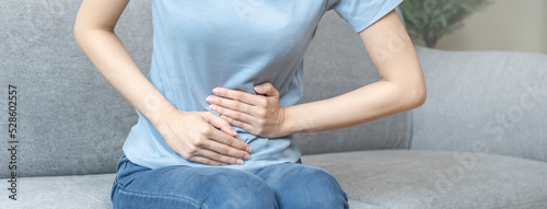 Flatulence asian young woman hand in stomach ache, suffer from food poisoning, abdominal pain and colon problem, gastritis or diarrhoea sitting on sofa at home. Patient belly, abdomen or inflammation.