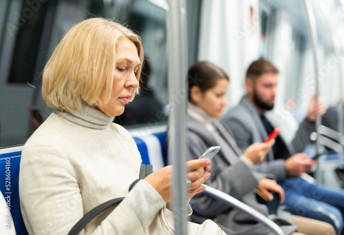 Adult woman browsing and typing messages on tablet in subway car leaning on handrail