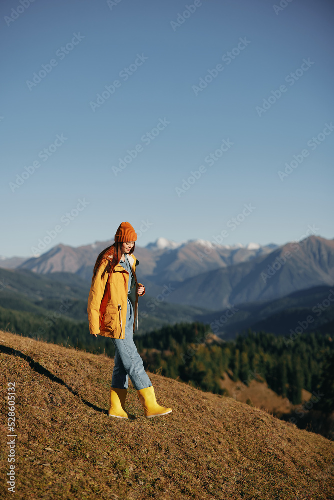 Woman autumn full-length walk on the hill smile with teeth and look at the mountains in a yellow raincoat and jeans happy hiking trip, freedom lifestyle Woman autumn full-length walk on the hill smile with teeth and look at the mountains in a yellow raincoat and jeans happy hiking trip, freedom lifestyle