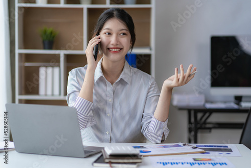 Asian woman working at home office on desk using calculator to calculate tax accounting expense report with graph document and laptop computer with smartphone.