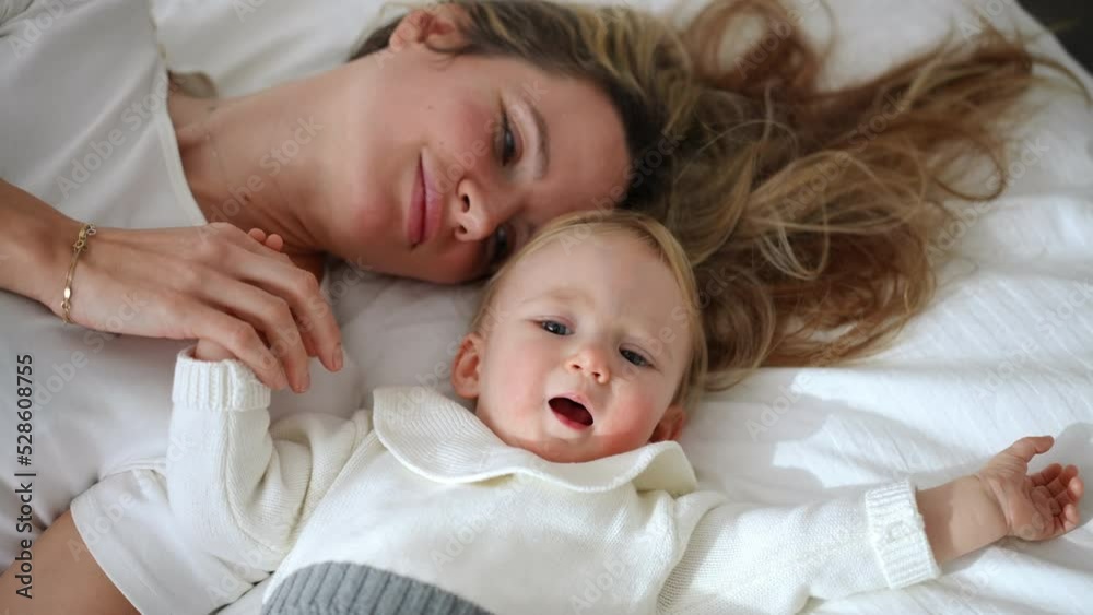 Curios little girl looking around and looking at camera as happy woman kissing child hand lying with baby on white soft cozy bed. High angle view portrait of Caucasian daughter and mother resting Curios little girl looking around and looking at camera as happy woman kissing child hand lying with baby on white soft cozy bed. High angle view portrait of Caucasian daughter and mother resting