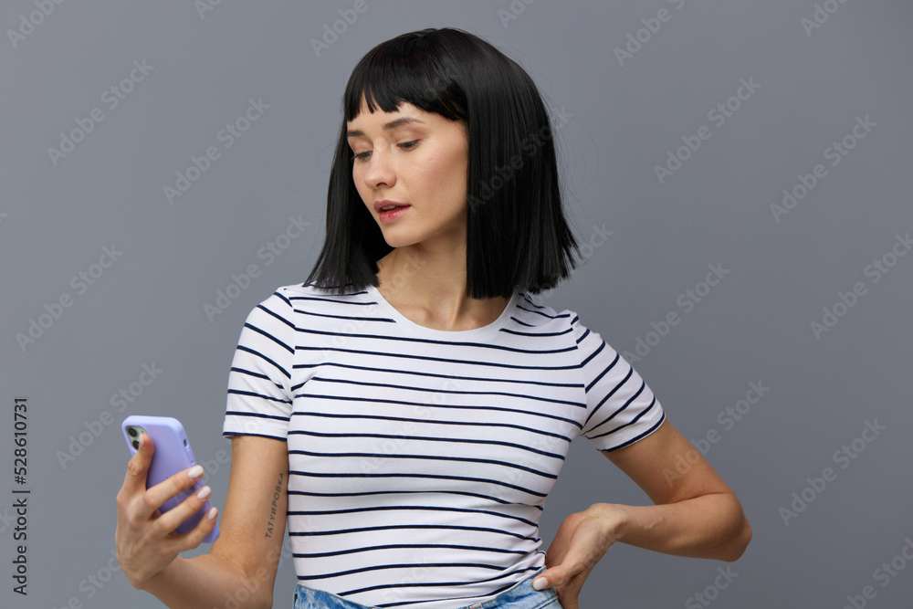 an attractive, beautiful woman with short black hair stands on a gray background in a striped cotton T-shirt and holds a smartphone in her hand looking at it. Horizontal photo with an empty space an attractive, beautiful woman with short black hair stands on a gray background in a striped cotton T-shirt and holds a smartphone in her hand looking at it. Horizontal photo with an empty space