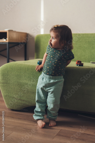 Full shot of a baby sucking a pacifier and looking at the bedroom window while leaning against the green sofa. Positive person. Beauty portrait. photo