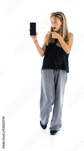 Surprised teen woman with mobile phone isolated on white background. Full length portrait of pretty girl pointing on smartphone. Stylish female in grey jeans and black t-shirt in studio.