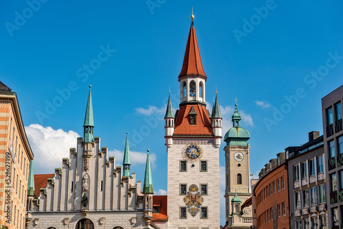 Der Turm des "Alten Rathaus" in der historischen Innenstadt von München bei sonnigem Sommerwetter und blauem Himmel mit dem Turm der "Heilig-Geist-Kirche" im Hintergrund