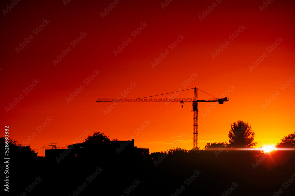 Tall crane silhouette against warm orange sunset sky. City skyline. Sun glow and flare. Dramatic rich saturated color.