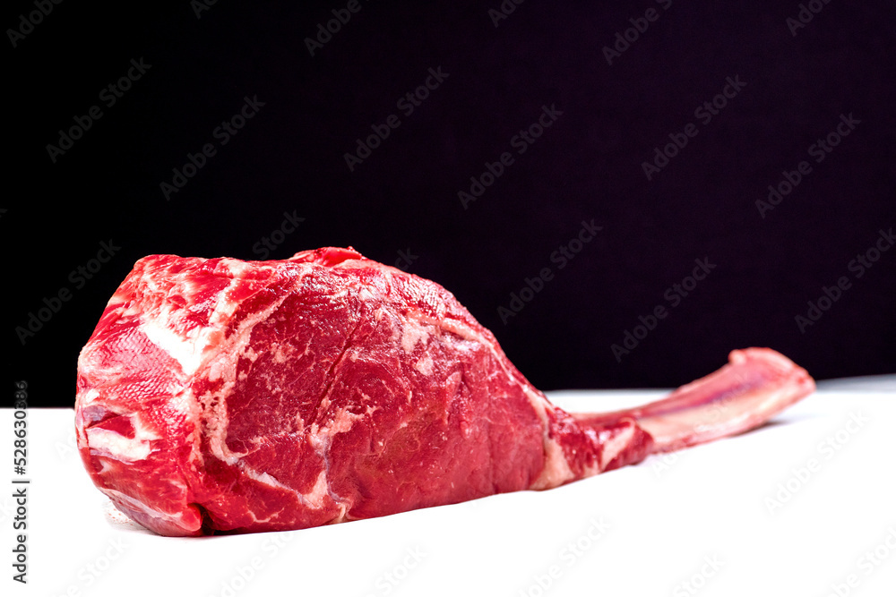 Obraz premium Fresh uncooked tomahawk steak on the bone on white cooking paper and black background. Prime cut of beef. High quality product with marbling and delicate flavor for special occasion. Butcher craft.