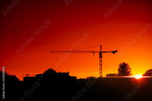 Tall crane silhouette against warm orange sunset sky. City skyline. Sun glow and flare. Dramatic rich saturated color.