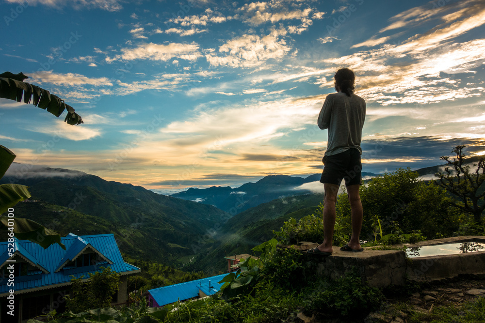 July 22nd 2021. Uttarakhand India. A man standing on a hill top looking at the landscape during sun set with beautiful multicolor cloudscape in the hills of the himalayas.