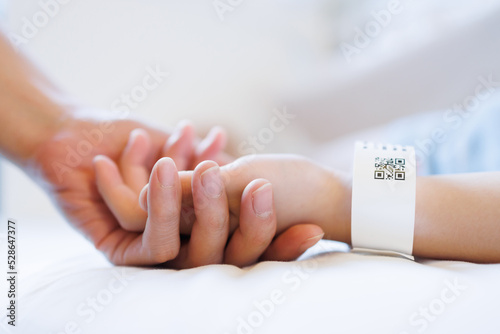 Foto father or mother hand hold kid patient hand with blured QR code patient tag on white bed in hospital room