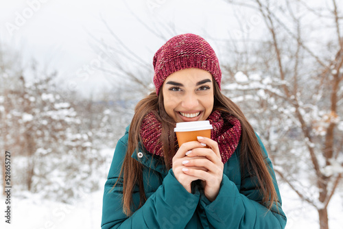 Pretty young woman in winter outfit holding disposable cup filled with hot coffee or tea. Girl holding mug of hot beverage in her hands and walks outdoors in winter.