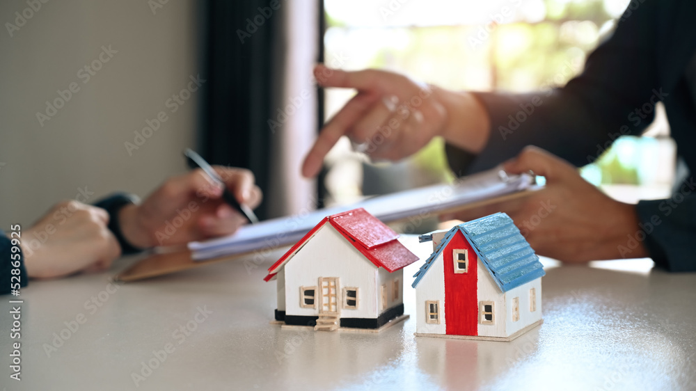Two small house models on white table with costumer signing contract with agent in background. Real estate and Property purchase concept Two small house models on white table with costumer signing contract with agent in background. Real estate and Property purchase concept