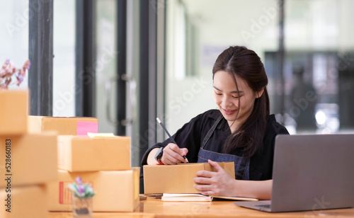Young beautiful happy asian business woman owner of SME online using laptop receive order from customer with parcel box packaging at her startup home office, online business seller and delivery © wichayada