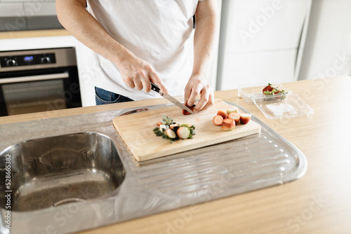 A man wearing a white t-shirt chopping up strawberries in a kitchen on a wooden board with a knife