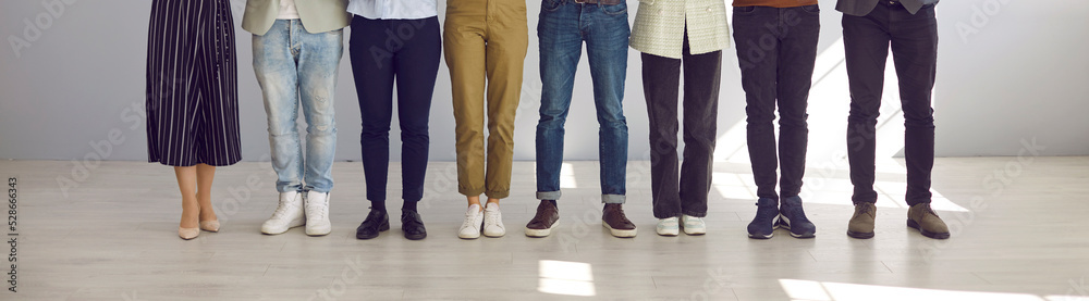 Cropped indoor low section shot of people's legs. Group of employees of different sexes and genders in pants and jeans standing in row on office floor. Banner. Smart and casual work dress code concept