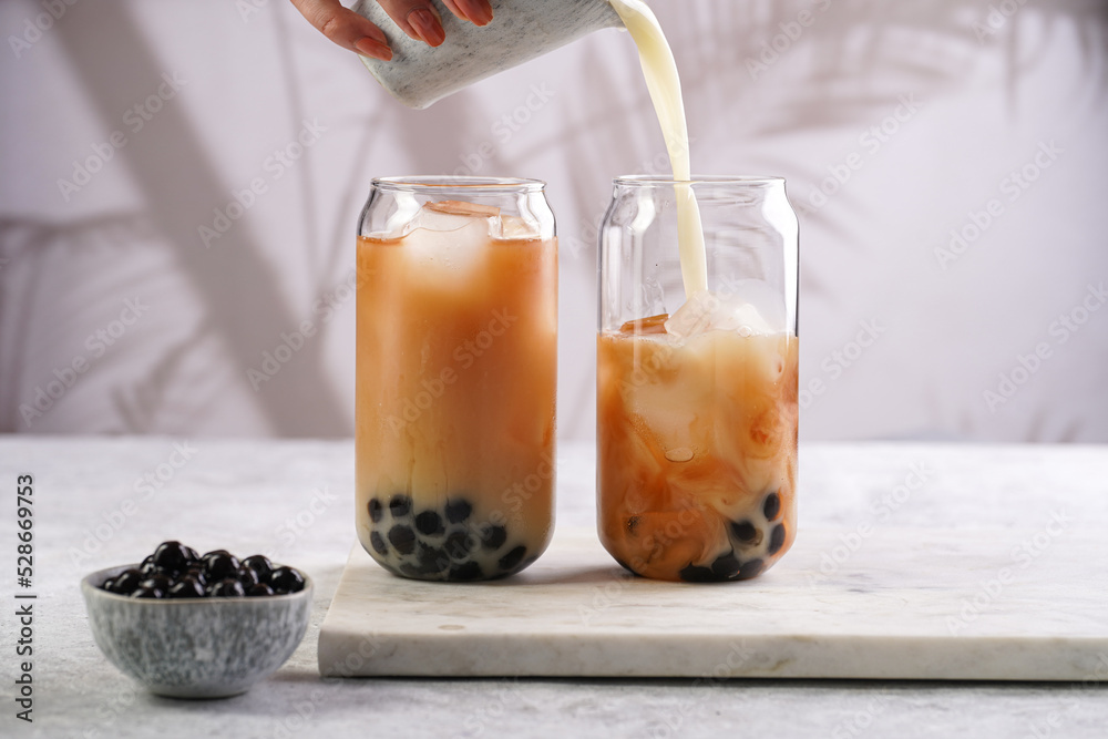 Female hand pouring milk into glass with black tea, ice cubes and cooked tapioca pearls for trendy bubble boba ice tea, two small grey ceramic bowls on marble board on pastel tropical background Female hand pouring milk into glass with black tea, ice cubes and cooked tapioca pearls for trendy bubble boba ice tea, two small grey ceramic bowls on marble board on pastel tropical background
