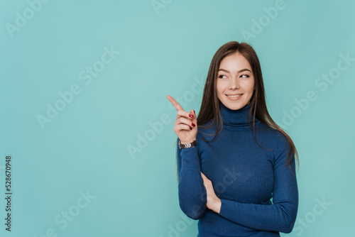 Fotobehang A beautiful European girl with long loose hair in a blue sweater points by  her index finger to the side, to an empty place, on a turquoise background looks at imaginary object