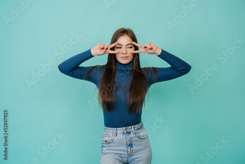 Gorgeous caucasian woman with long loose hair in blue sweater and blue jeans posing at studio over turquoise backdrop making victory sign near eyes by both hands. Mockup beauty and fashion.