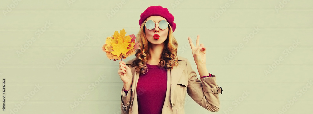 Autumn portrait of beautiful young woman model with yellow maple leaves blowing her lips with lipstick wearing pink french beret on gray background Autumn portrait of beautiful young woman model with yellow maple leaves blowing her lips with lipstick wearing pink french beret on gray background