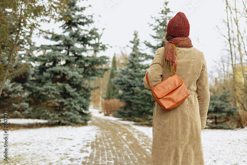 Back view of woman in warm coat and hat walking on snowy pathway in winter park in city 