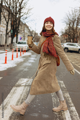 Side view full body of carefree woman in coat and warm hat with scarf and blank cup of takeaway coffee crossing road on crosswalk in city in winter 