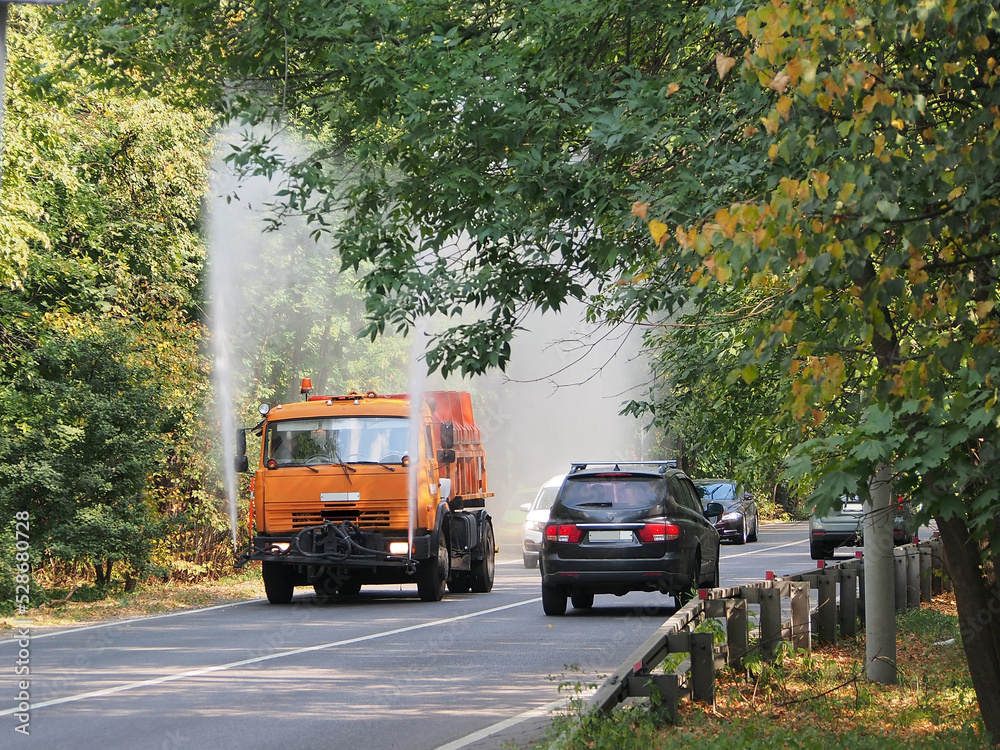 Obraz premium Watering the streets with an orange car with a tank, a hot summer day. The work of municipal equipment on city roads.
