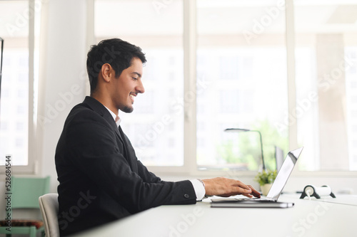 Optimistic hispanic male office employee using laptop in modern coworking space, businessman in formal suit working on computer, watching at the screen and smiling, side view