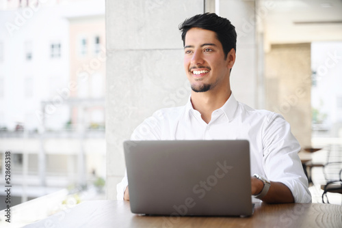 Handsome young dark-haired latin man in white smart casual shirt working on a laptop on the terrace of summer cafe during break. Developer, programmer, copywriter enjoying remote work outdoors