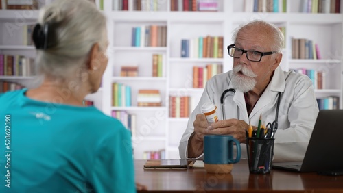 Mature senior male doctor discussing treatment with prescription drug to female patient in modern clinic with books background.