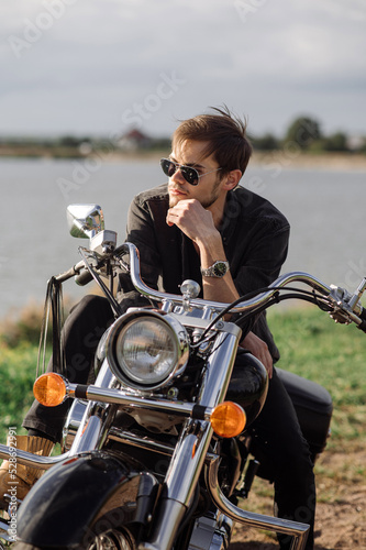 Young handsome male biker is sitting on his motor bike outdoors at the lake background © rostyslav84