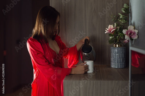 Pour hot water glass tea, Woman making morning tea to have with breakfast at home, A girl in a satin nightgown and red robe making tea in the kitchen photo