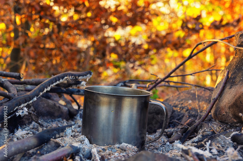 A metal tourist mug is heated in the coals of a burnt out fire against a background of blurry orange foliage. Scene from the autumn hike