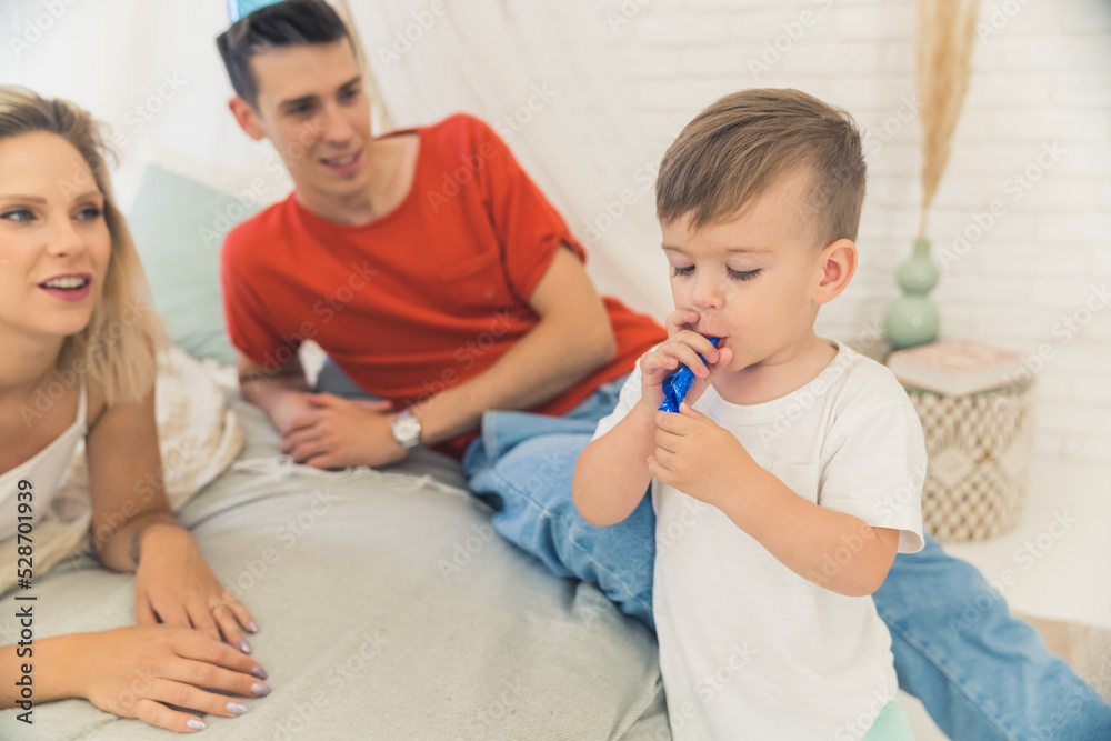 Fototapeta premium Party horn experience for the first time in life. Young caucasian couple watching their son trying to blow dark blue party horn. Birthday concept. High quality photo