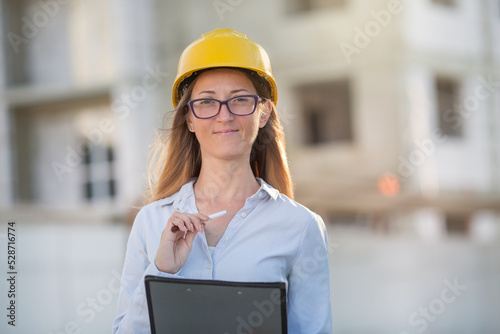 a young blonde woman is standing at a construction site with documents. She is wearing glasses, a light blouse, a yellow helmet.. © Olga