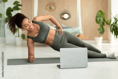 Fit african american woman standing in side plank position on yoga mat, training with laptop computer at home