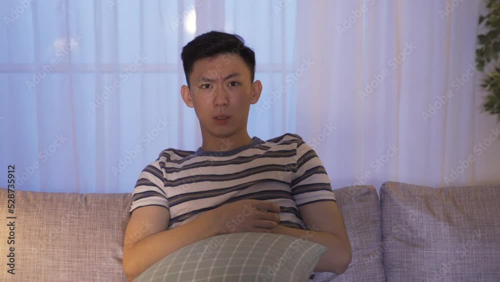 front portrait shot frightened Asian male is touching his chest to calm down while watching a scary movie on the couch with a pillow in the living room at home. front portrait shot frightened Asian male is touching his chest to calm down while watching a scary movie on the couch with a pillow in the living room at home.