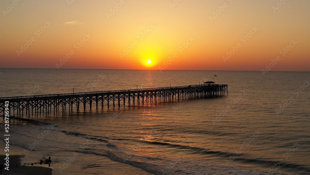 Sun rising over peaceful ocean with fishing pier on the beach