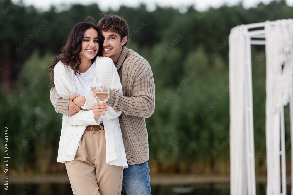 Smiling couple in cardigans hugging and holding wine outdoors
