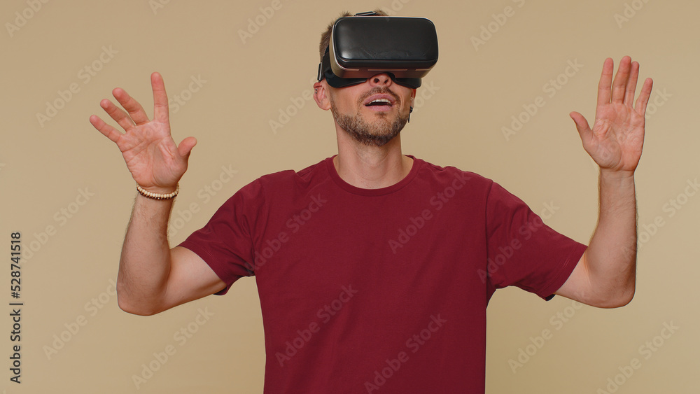 Bearded handsome young man using headset helmet app to play simulation drawing game. Watching virtual reality 3D 360 video. Adult male guy in VR goggles isolated on beige studio background indoors Bearded handsome young man using headset helmet app to play simulation drawing game. Watching virtual reality 3D 360 video. Adult male guy in VR goggles isolated on beige studio background indoors