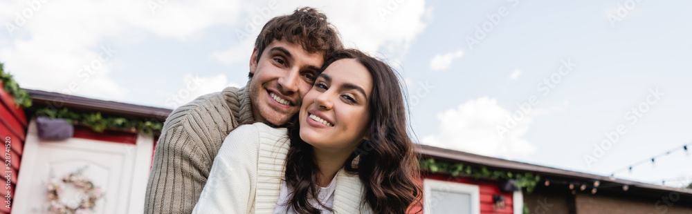 Smiling couple in knitted cardigans looking at camera near blurred house outdoors, banner