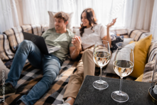 Glasses of wine near blurred couple with laptop on bed in camper