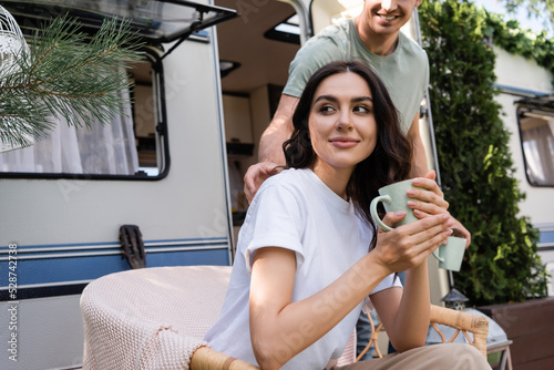 Smiling woman holding cup while sitting on armchair near boyfriend and camper van outdoors