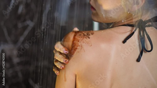Young woman puts anti-cellulite dark coffee scrub on soft wet skin of shoulder making massage with hand in shower cabin at home slow motion closeup