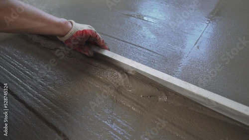 A builder in protective gloves pours a self-levelling screed onto the floor and applies a liquid cement mortar with a metal spatula. Leveling the mortar on the floor using metal guides. The master photo