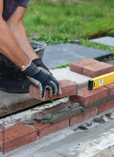 Male hands in protective gloves holding red bricks. Construction worker builds masonry. Brickwork close up photo. 