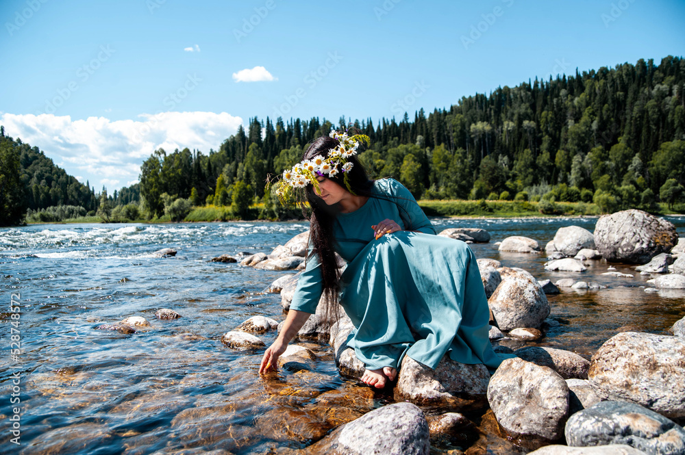 A beautiful young girl of Middle Eastern appearance in a turquoise dress and with a beautiful wreath of white daisies and ferns, wearing black long hair, came to a clear lake and sat on a stone
