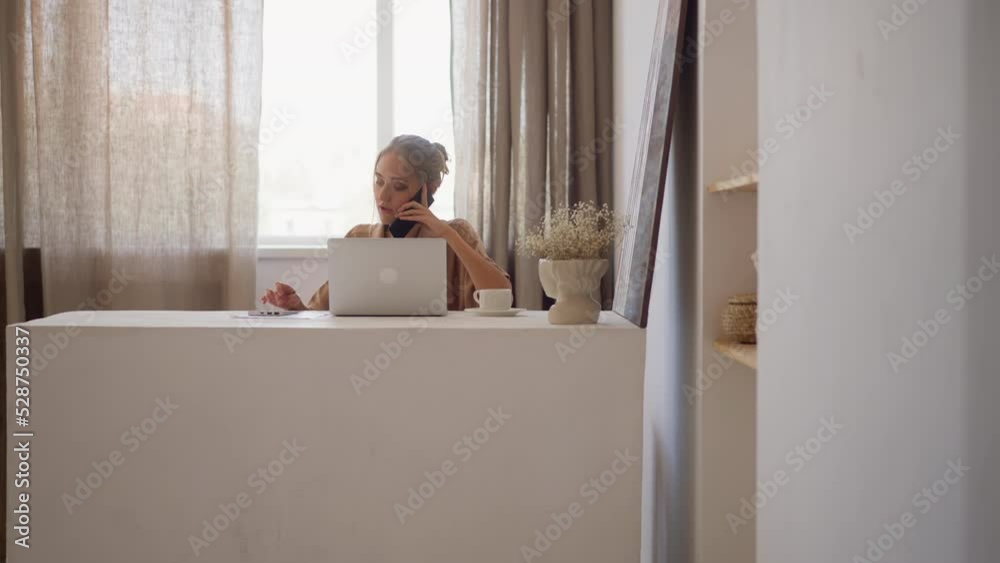 Blonde woman in stylish beige shirt works on notebook at white table talking on mobile phone with colleague against bright window in office room slow motion Blonde woman in stylish beige shirt works on notebook at white table talking on mobile phone with colleague against bright window in office room slow motion